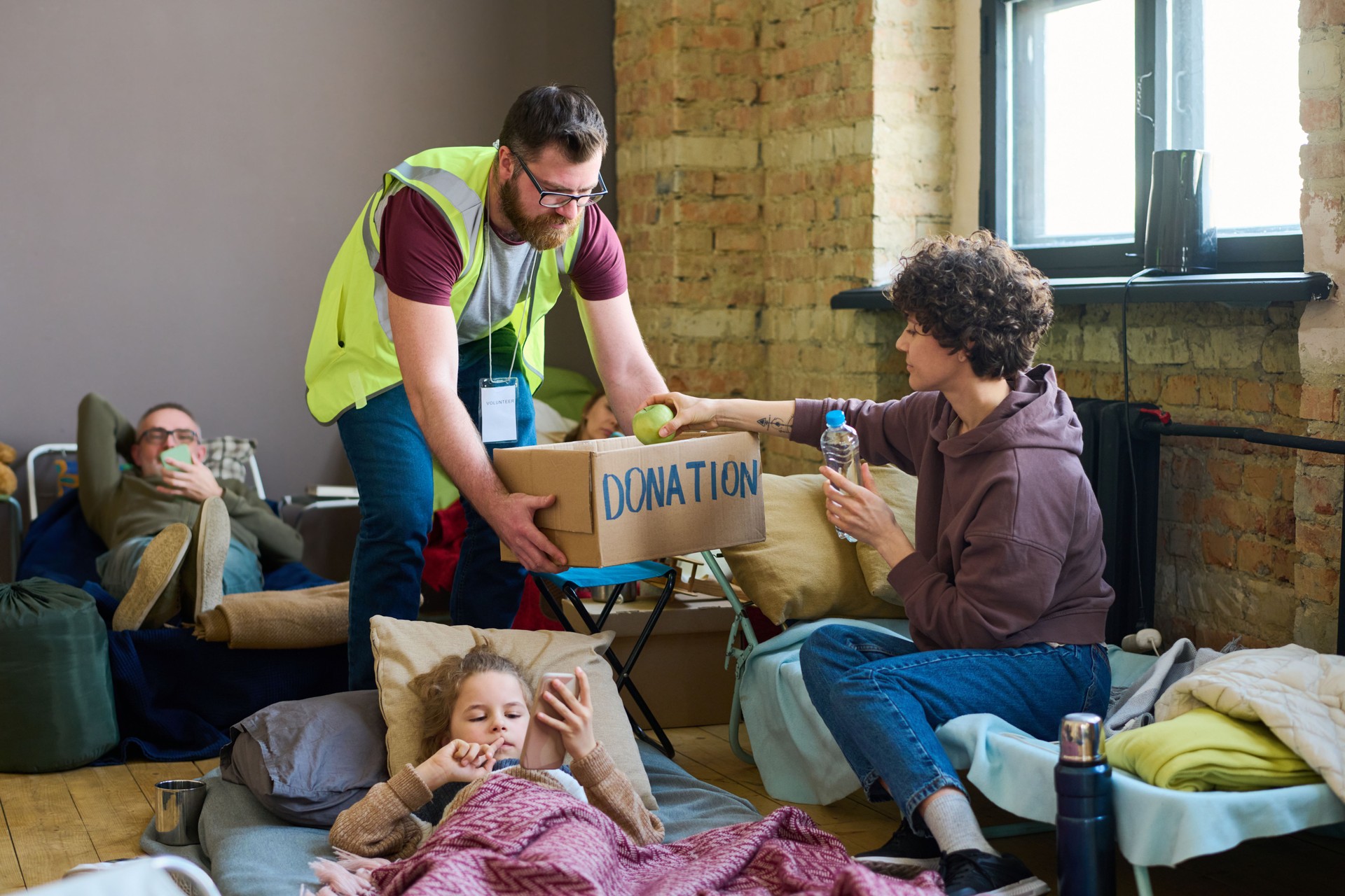 Young male volunteer with donation box bending in front of female refugee